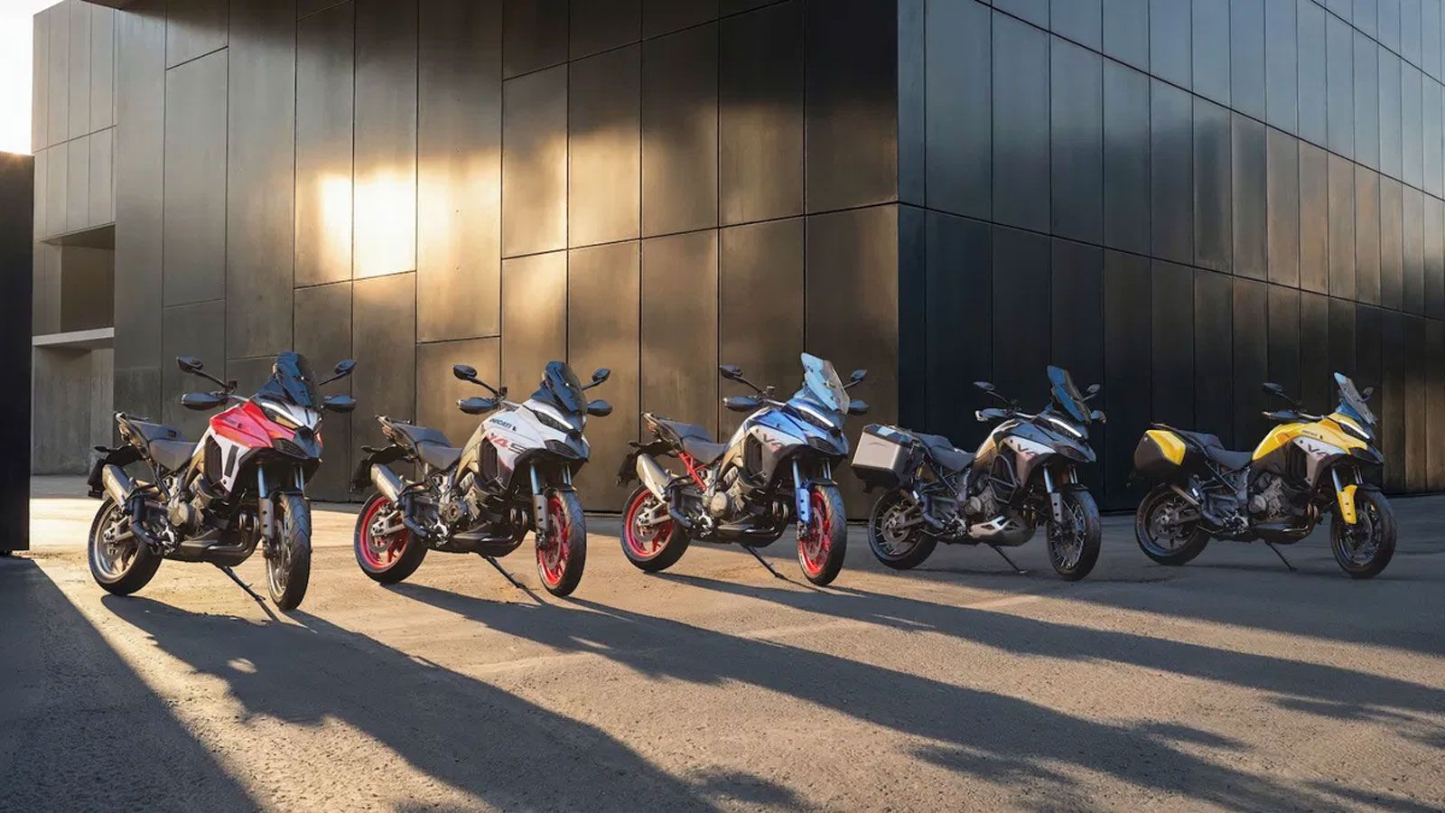 Πέντε Ducati Multistrada V4 σε διαφορετικές ειδικές βαφές και εκδόσεις, παρουσιασμένες σε ανοιχτό χώρο με ηλιοβασίλεμα, στο πλαίσιο του Ducati Factory