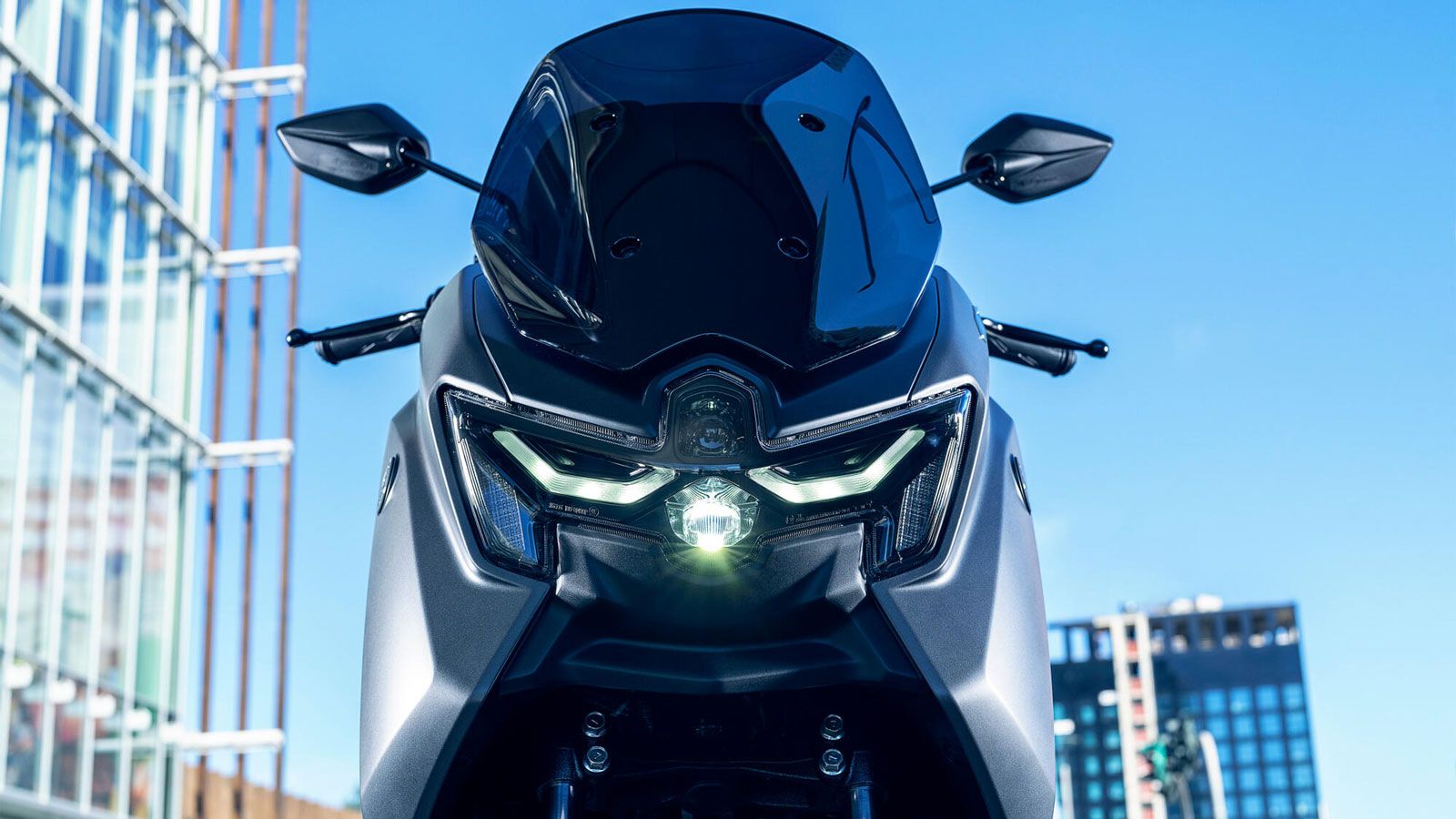 Μπροστινή όψη Yamaha NMAX 155 Tech MAX με LED προβολέα και ψηλή ζελατίνα, σε αστικό περιβάλλον με σύγχρονα κτίρια στο φόντο.
